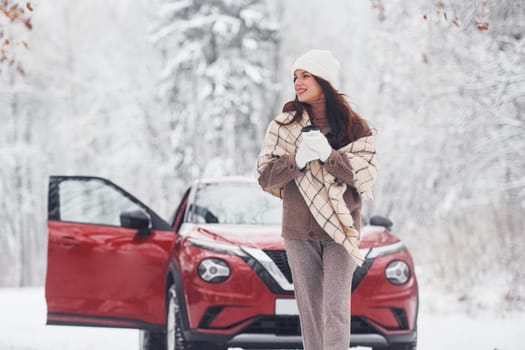 In the forest. Beautiful young woman is outdoors near her red automobile at winter time.
