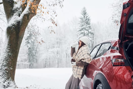 In the forest. Beautiful young woman is outdoors near her red automobile at winter time.