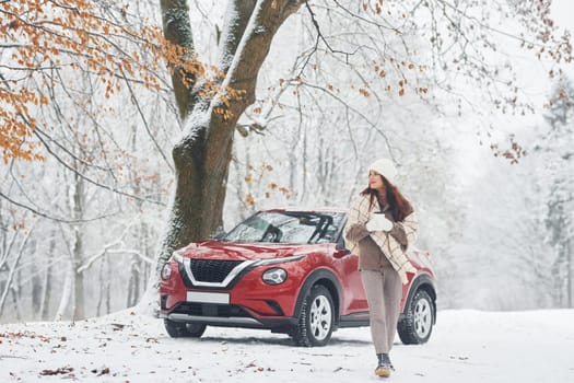 Rural scene. Beautiful young woman is outdoors near her red automobile at winter time.