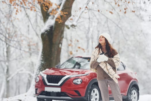 Rural scene. Beautiful young woman is outdoors near her red automobile at winter time.