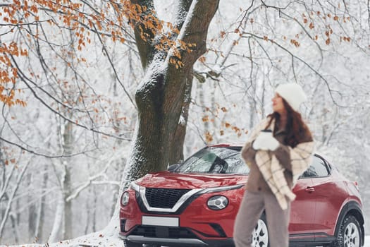 Rural scene. Beautiful young woman is outdoors near her red automobile at winter time.
