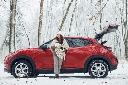 Rural scene. Beautiful young woman is outdoors near her red automobile at winter time.