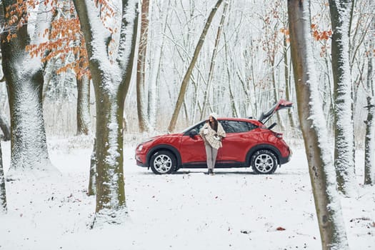 Rural scene. Beautiful young woman is outdoors near her red automobile at winter time.
