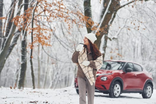 Trees are covered with snow. Beautiful young woman is outdoors near her red automobile at winter time.