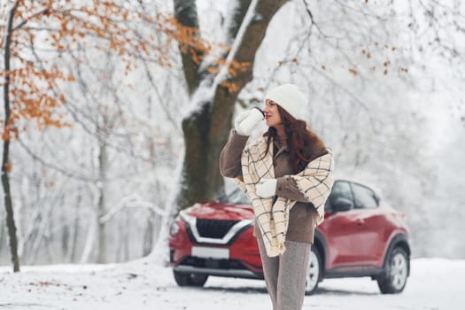 Trees are covered with snow. Beautiful young woman is outdoors near her red automobile at winter time.