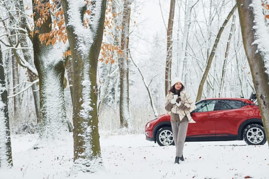 Trees are covered with snow. Beautiful young woman is outdoors near her red automobile at winter time.