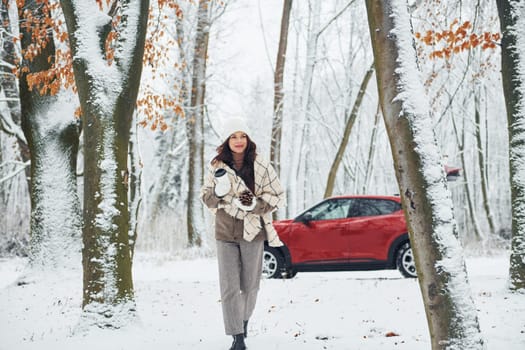 Trees are covered with snow. Beautiful young woman is outdoors near her red automobile at winter time.