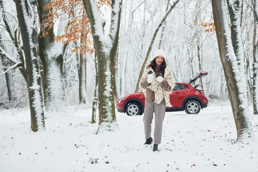 Having a walk in the forest. Beautiful young woman is outdoors near her red automobile at winter time.