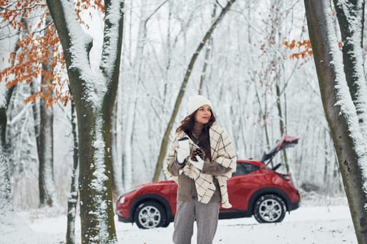 Having a walk in the forest. Beautiful young woman is outdoors near her red automobile at winter time.