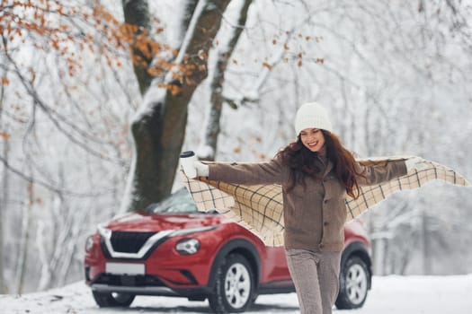 Having a walk in the forest. Beautiful young woman is outdoors near her red automobile at winter time.