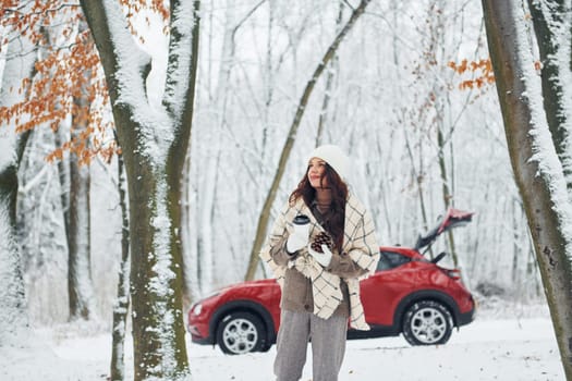 Having a walk in the forest. Beautiful young woman is outdoors near her red automobile at winter time.