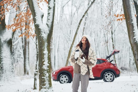 Having a walk in the forest. Beautiful young woman is outdoors near her red automobile at winter time.