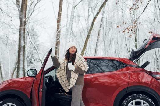 New car is in the forest. Beautiful young woman is outdoors near her red automobile at winter time.
