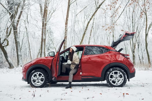 New car is in the forest. Beautiful young woman is outdoors near her red automobile at winter time.