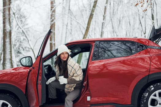 New car is in the forest. Beautiful young woman is outdoors near her red automobile at winter time.