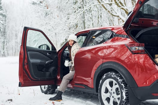 New car is in the forest. Beautiful young woman is outdoors near her red automobile at winter time.