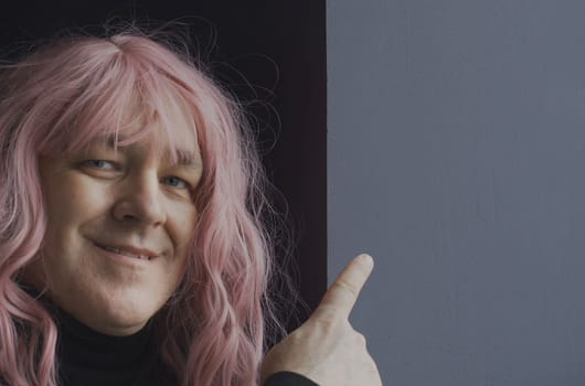Portrait of a man in a pink wig, looks and points a blank space for text with his finger. close-up.