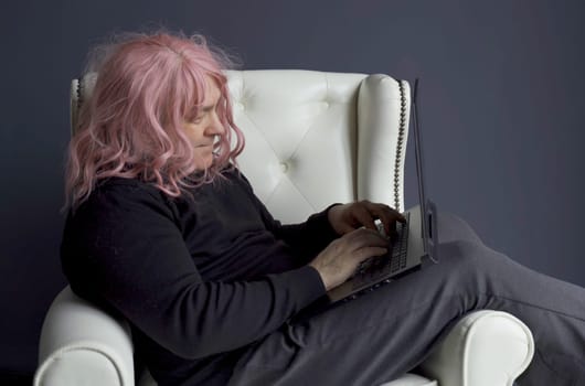 A man in a pink wig sits in a white chair and works at a computer.