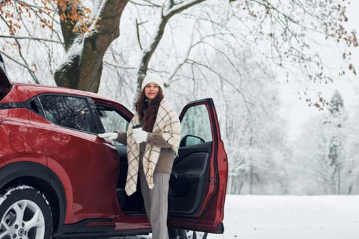 Beautiful young woman is outdoors near her red automobile at winter time.