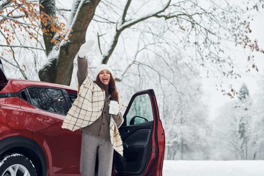 Beautiful young woman is outdoors near her red automobile at winter time.