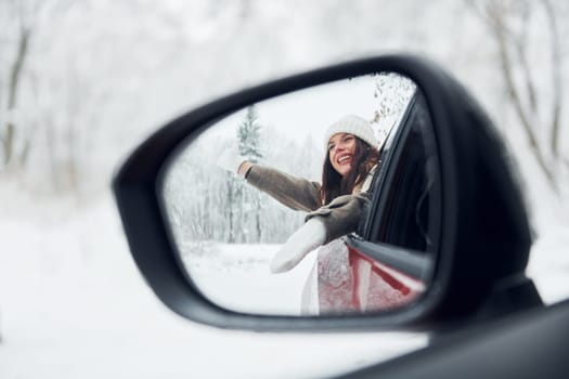 View in the side mirror. Beautiful young woman is outdoors near her red automobile at winter time.