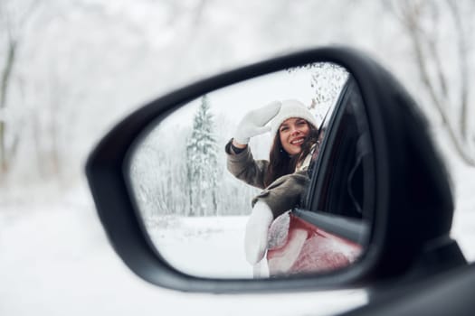 View in the side mirror. Beautiful young woman is outdoors near her red automobile at winter time.