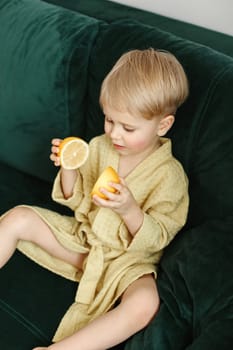 A boy in a yellow bathrobe is sitting on a green sofa, playing with halves of an orange. Vertical frame. View from above