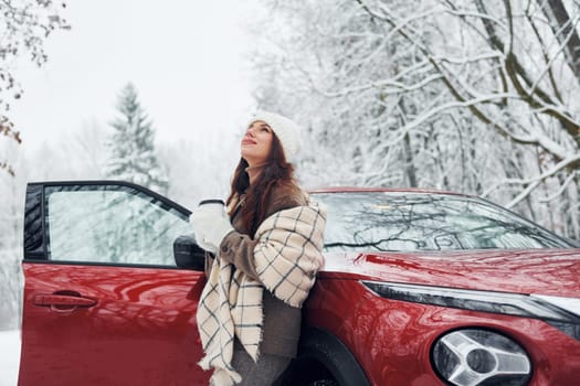 Non urban scene. Beautiful young woman is outdoors near her red automobile at winter time.