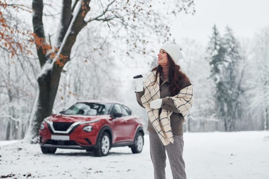 Non urban scene. Beautiful young woman is outdoors near her red automobile at winter time.