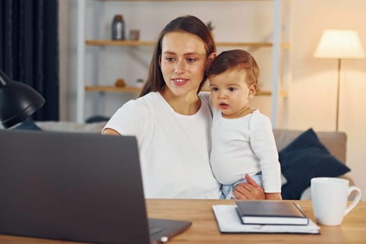 Sitting by the table with laptop. Mother with her little daughter is at home together.