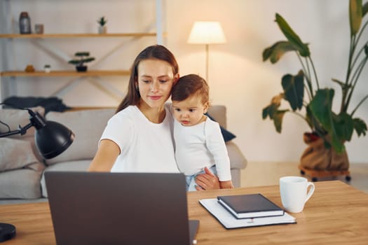 Sitting by the table with laptop. Mother with her little daughter is at home together.