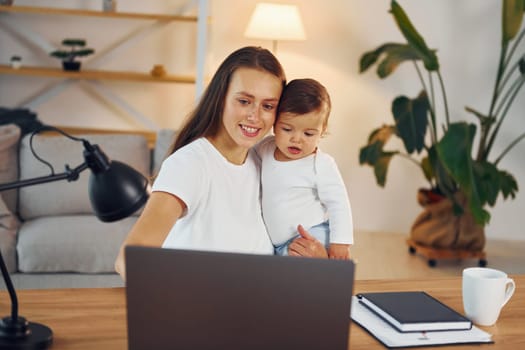 Sitting by the table with laptop. Mother with her little daughter is at home together.