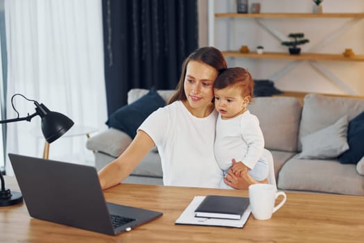 Sitting by the table with laptop. Mother with her little daughter is at home together.