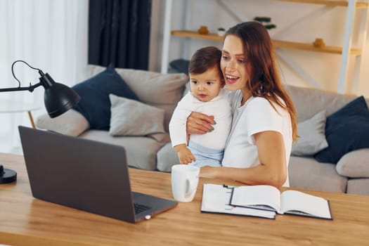 Laptop is on the table. Mother with her little daughter is at home together.