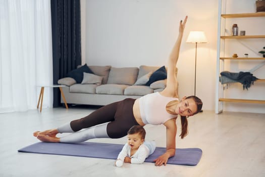 Woman doing exercises. Mother with her little daughter is at home together.