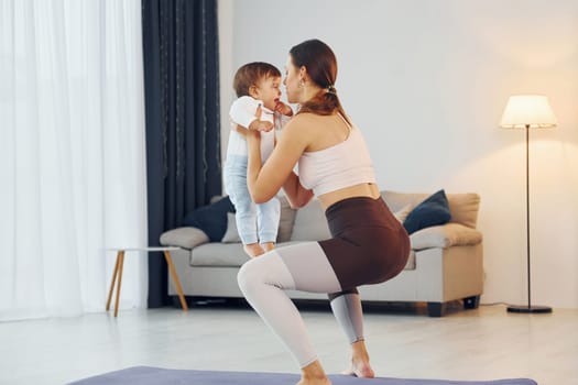 Standing on the mat and doing exerises. Mother with her little daughter is at home together.