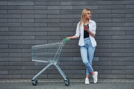 Beautiful blonde in casual clothes with shopping cart is outdoors at sunny daytime.