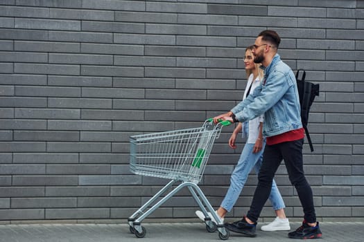 With shopping cart. Young stylish man with woman in casual clothes outdoors together. Conception of friendship or relationships.