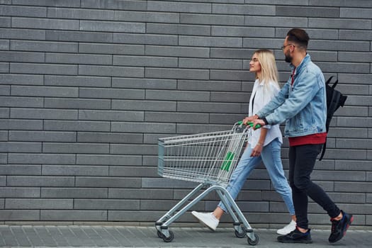 With shopping cart. Young stylish man with woman in casual clothes outdoors together. Conception of friendship or relationships.