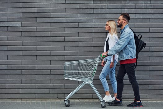 With shopping cart. Young stylish man with woman in casual clothes outdoors together. Conception of friendship or relationships.