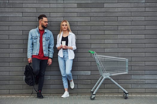 With shopping cart. Young stylish man with woman in casual clothes outdoors together. Conception of friendship or relationships.