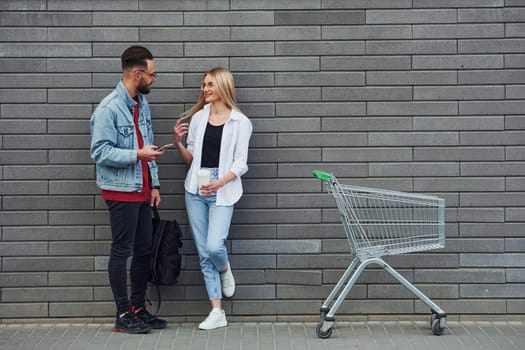 With shopping cart. Young stylish man with woman in casual clothes outdoors together. Conception of friendship or relationships.