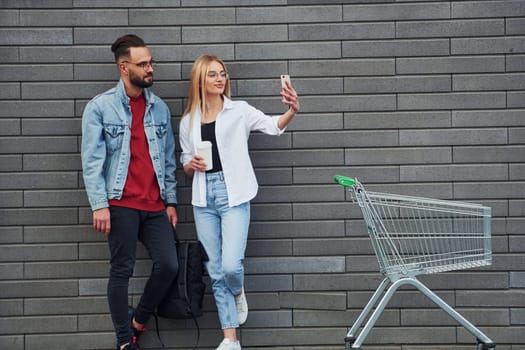 With shopping cart. Young stylish man with woman in casual clothes outdoors together. Conception of friendship or relationships.