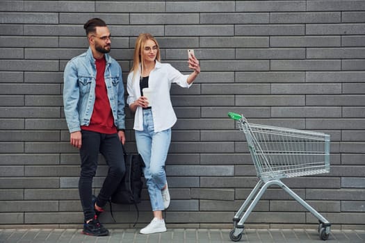 With shopping cart. Young stylish man with woman in casual clothes outdoors together. Conception of friendship or relationships.