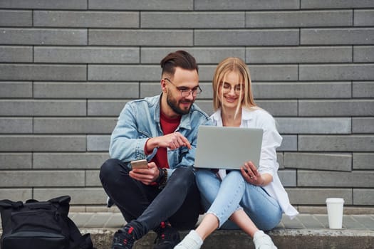 Using laptop. Young stylish man with woman in casual clothes sitting outdoors together. Conception of friendship or relationships.