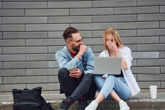 Using laptop. Young stylish man with woman in casual clothes sitting outdoors together. Conception of friendship or relationships.