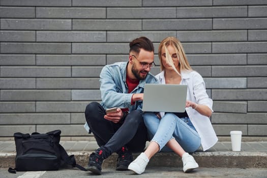 Using laptop. Young stylish man with woman in casual clothes sitting outdoors together. Conception of friendship or relationships.