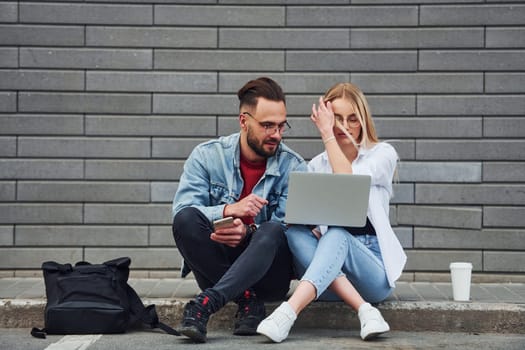 Using laptop. Young stylish man with woman in casual clothes sitting outdoors together. Conception of friendship or relationships.