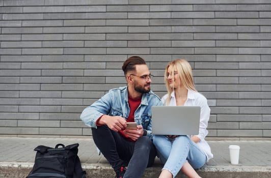 Using laptop. Young stylish man with woman in casual clothes sitting outdoors together. Conception of friendship or relationships.