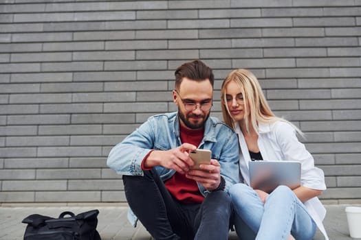 Using laptop. Young stylish man with woman in casual clothes sitting outdoors together. Conception of friendship or relationships.